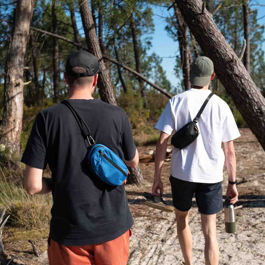 Two people walking through a pine forest wearing sling bags